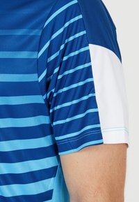 Blå idrottströja med horisontella ränder i olika nyanser av blått, med en vit axelpanel och ett slätt, lätt tyg.