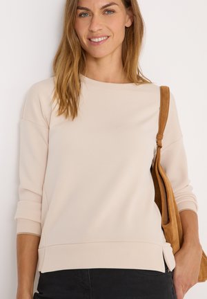 Sweatshirt beige clair avec des manches longues, un col rond et des fentes sur les côtés, associé à un sac en daim brun sur un fond neutre.
