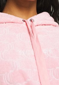 Rosa Kapuzenpullover aus Frottee mit erhabenem Logomuster, Kordelzug mit silbernen Akzenten und aufgesticktem "Juicy Couture" Schriftzug auf der Vorderseite.