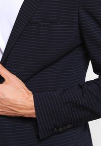 Man justerar en marinblå kostymjacka med smala ränder över en vit skjorta, visar hand och ärm med fyra knappar.