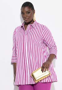 Abito camicia a righe rosa e bianche, in tessuto leggero, con colletto e maniche a tre quarti. In mano tiene una clutch oro lucido. Pantaloni aderenti.