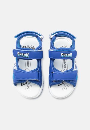 Kék nyitott orrú gyerek szandálok állítható pántokkal, amelyeken „SHARK” felirat szerepel, fehér talpak cápa grafikával belül, egyszerű háttéren bemutatva.