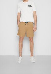 Bañador color beige con cintura elástica y cordón azul, combinado con una camiseta gráfica blanca con texto verde, llevado con zapatillas blancas.