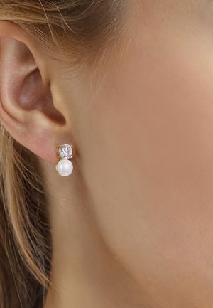 Oreja con pendiente de botón de oro con una piedra preciosa redonda y transparente sobre una perla blanca.