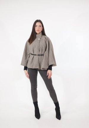 Femme aux longs cheveux foncés portant une cape en laine grise ceinturée, un haut à manches longues noires, un pantalon skinny gris et des bottines noires, debout sur un fond blanc.