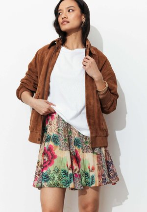 Femme portant une veste en daim marron sur une chemise blanche et une jupe colorée à motif floral, posant devant un fond blanc.