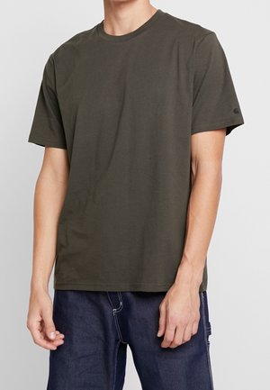 Mężczyzna ubrany w oliwkowozielony t-shirt z krótkim rękawem i ciemnoniebieskie dżinsy, stojący na tle jednolitego jasnego tła.