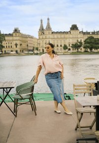 Blouse collier à manches courtes rose clair ; jupe en denim avec une fente à l'avant ; ballerines beiges. Cadre de café extérieur près de l'eau avec des accents verts.