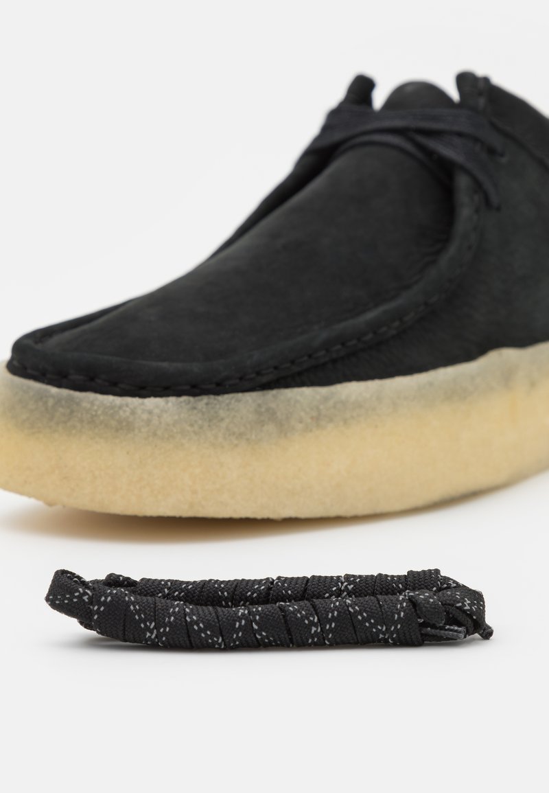 Scarpa in suede nera con un design morbido e arrotondato e una suola in gomma leggera color tan, leggermente consumata. Include un laccio in tessuto nero con texture.