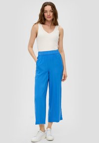 Witte geribde tanktop, losse felblauwe broek met elastische tailleband, model draagt witte sneakers en staat op een grijze achtergrond.