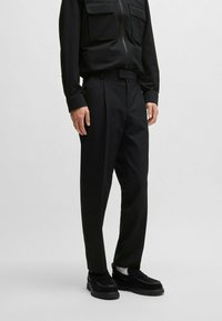 Schwarze Hosen mit einem maßgeschneiderten Design, featuring eine glatte Vorderseite, Taschen und eine glatte Textur, kombiniert mit schwarzen Slippern für einen stimmigen Look.