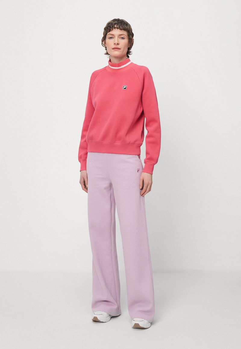 Felpa rosa con scollatura a coste e patch con logo, abbinata a pantaloni larghi lilla. Sneakers bianche completano l'outfit.