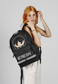 Mochila negra con un personaje de dibujos animados y el texto "MIÉRCOLES". Presenta correas ajustables, un bolsillo delantero y acentos en rayas blancas.