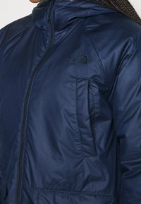 Marineblauwe geïsoleerde jas met een ritssluiting aan de voorkant, een capuchon en een logo. Gemaakt van glad, duurzaam materiaal met diagonale textuur en zakken.