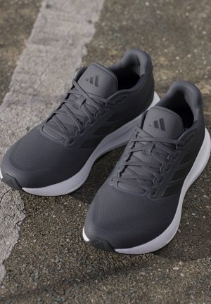 Paire de baskets de sport noires avec semelles blanches, posées sur l'asphalte près d'une ligne blanche effacée.