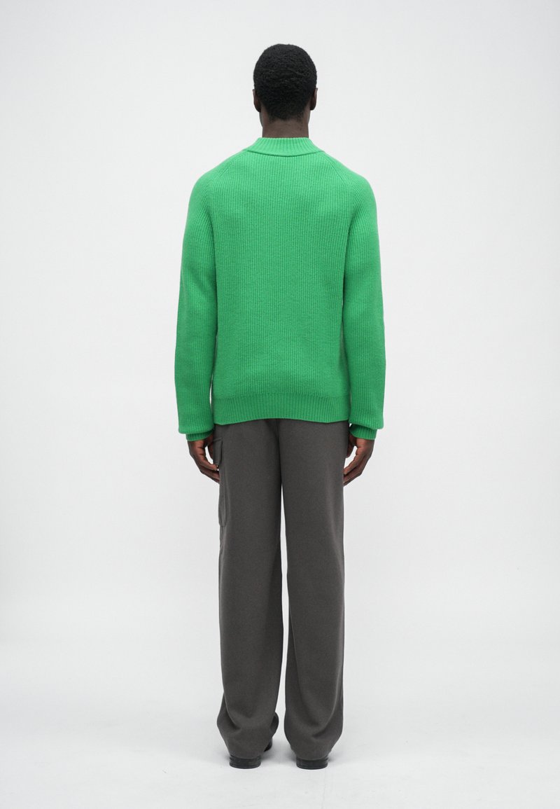 Pull en laine vert avec une texture côtelée, encolure ronde et dos droit, associé à un pantalon gris ample.