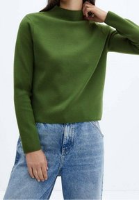 Pull en maille verte avec un col montant et des manches longues, assorti à un jean en denim bleu clair. Le tissu a une texture lisse.