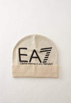 Bonnet tricoté beige avec le logo noir "EA7 Emporio Armani" imprimé sur le devant, posé à plat sur une surface blanche.