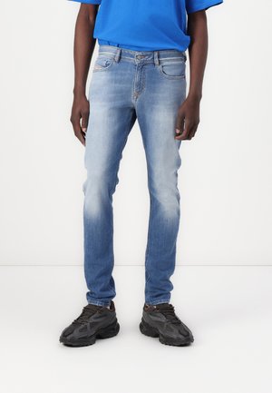Personne portant un jean skinny bleu délavé, des chaussures de sport noires et un t-shirt bleu à manches courtes, debout devant un fond blanc.