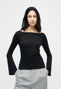 Γυναίκα με σκουρόχρωμα μαλλιά μέχρι τους ώμους, που φοράει μαύρο μακρυμάνικο τοπ και ανοιχτό γκρι παντελόνι με λεπτές ρίγες, έχοντας τα χέρια στις τσέπες.