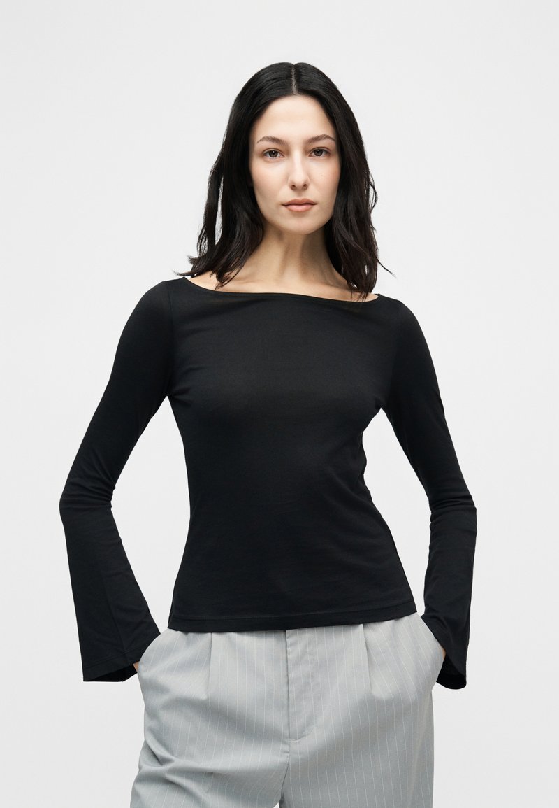 Γυναίκα με σκουρόχρωμα μαλλιά μέχρι τους ώμους, που φοράει μαύρο μακρυμάνικο τοπ και ανοιχτό γκρι παντελόνι με λεπτές ρίγες, έχοντας τα χέρια στις τσέπες.