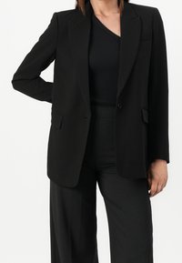 Schwarzer, taillierter Blazer aus glattem Stoff mit Reverskragen, Ein-Knopf-Verschluss und zwei Fronttaschen. Über einem schwarzen Oberteil gestylt.