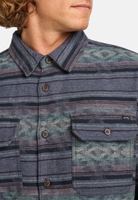 Chemise à boutons en tissu texturé avec des rayures bleu foncé et clair, accentuées de vert et de violet, deux poches poitrine et un col pointu.