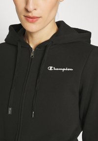 Felpa nera con zip realizzata in tessuto morbido, con cappuccio, cordini e un logo "Champion" in argento sul petto.