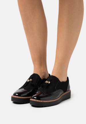 Chaussures en suede noir et cuir verni avec des accents de glands et des détails de brogue, présentant une semelle de plateforme épaisse et texturée.