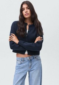 Ung kvinna med lång mörk hår som bär en marinblå knäppt cardigan och ljusblå jeans, stående med armarna i kors mot en enfärgad bakgrund.