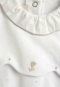 Top in tessuto bianco con colletto a volant, decorato con piccole perline dorate e una rosa ricamata; presenta una chiusura a bottoni al collo.