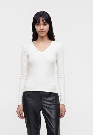 Junge Frau mit langen schwarzen Haaren, die ein weißes, langärmliges V-Ausschnitt-Shirt und schwarze Lederhosen trägt, steht vor einem schlichten Hintergrund.