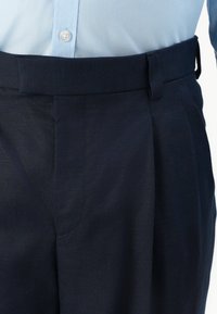 Mann trägt marineblaue, gefaltete Hose mit breitem Bund und hellblauem, in die Hose gestecktem Hemd.