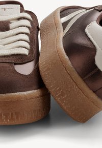 Baskets marron avec des lacets blancs et des bandes latérales, dotées de semelles en gomme texturées, présentées en gros plan sous des angles arrière et latéral.