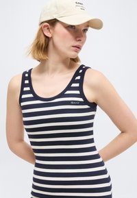 Γυναίκα που φοράει μπλε σκούρο και λευκό ριγέ αμάνικο τοπ και μπεζ καπέλο του μπέιζμπολ, κοιτάζοντας πλάγια με τα χέρια πίσω από την πλάτη της.