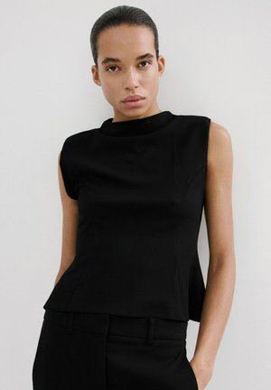 Ženska z zaglajenimi lasmi, oblečena v brezrokavnik in črne hlače, na preprostem belem ozadju.