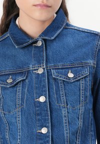 Jaqueta de denim azul com um colarinho clássico, fechos de botão prateados e dois bolsos no peito. Apresenta costuras visíveis e uma textura suave.