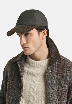 Dark gray cap with ridged brown brim, featuring embroidered brand logo. Model wears a thick cable-knit sweater and a textured tweed coat.