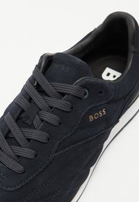 Tengerészkék velúr sneaker, texturált fűzésekkel, domború "BOSS" logóval és fehér gumitalppal, amelynek mintázata van.