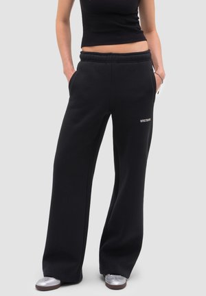 Kobieta ubrana w czarne, szerokie spodnie dresowe z logo WRSTBHVR, czarny krótki top na ramiączkach i srebrne buty sportowe, z rękami w kieszeniach.