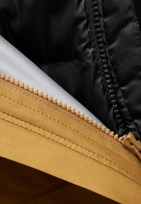 La veste zippée présente une coque extérieure brun doré, une doublure intérieure noire et une couche intérieure argentée, mettant en valeur des textures et des couleurs contrastées.