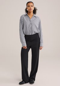 Črno-bela prugasta srajca z gumbom in ovratnikom, v kombinaciji s črnimi širokimi hlačami in črnimi čevlji. Gladek tekstilni videz.