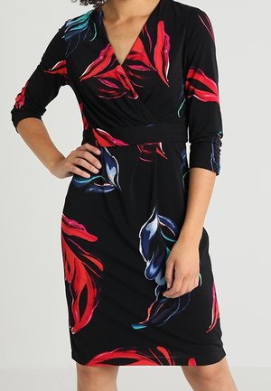 Mujer con vestido negro hasta la rodilla con patrones florales abstractos en rojo, azul y verde azulado, y mangas tres cuartos.