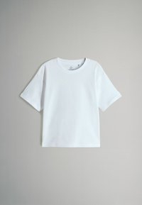 Biała, krótko rękawowa koszulka typu crewneck wisząca na tle neutralnym w jasnoszarym kolorze.