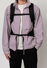 Personne portant une veste zippée violet clair, une chemise blanche, un sac à dos noir avec des sangles poitrine et taille, et un pantalon noir.