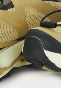 Zapatilla deportiva en tonos marrón y negro, con materiales de malla y sólidos, que presenta un patrón texturizado y una suela flexible. Incluye un detalle de bloqueo de cordones en negro.