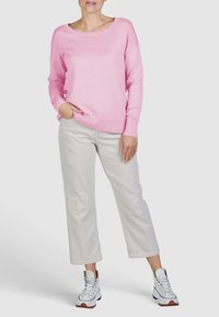 Vrouw in een lichtroze trui met lange mouwen, lichtgrijze cropped broek met kleine slijtage details, en witte hoge sneakers die staat.