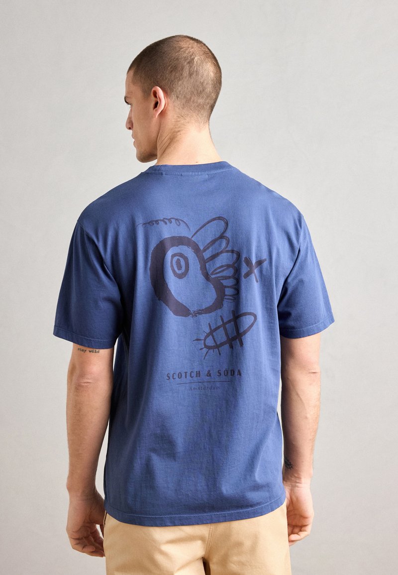 Marineblauwe katoenen t-shirt met een grafisch ontwerp op de achterkant met abstracte vormen en de tekst "SCOTCH & SODA" aan de onderkant. Korte mouwen.