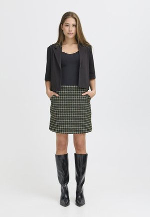 Jeune femme portant un blazer court noir, un haut noir, une jupe à carreaux verts et blancs, et des bottes noires montantes, se tenant avec les mains dans les poches.