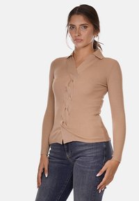 Maglione in maglia a coste beige con scollo a V, caratterizzato da un design attorcigliato sui lati e maniche lunghe. Abbinato a jeans scuri.